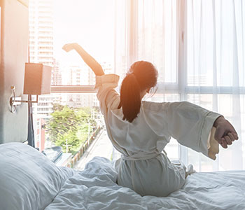 Lady sitting on bed facing rising sun and streching